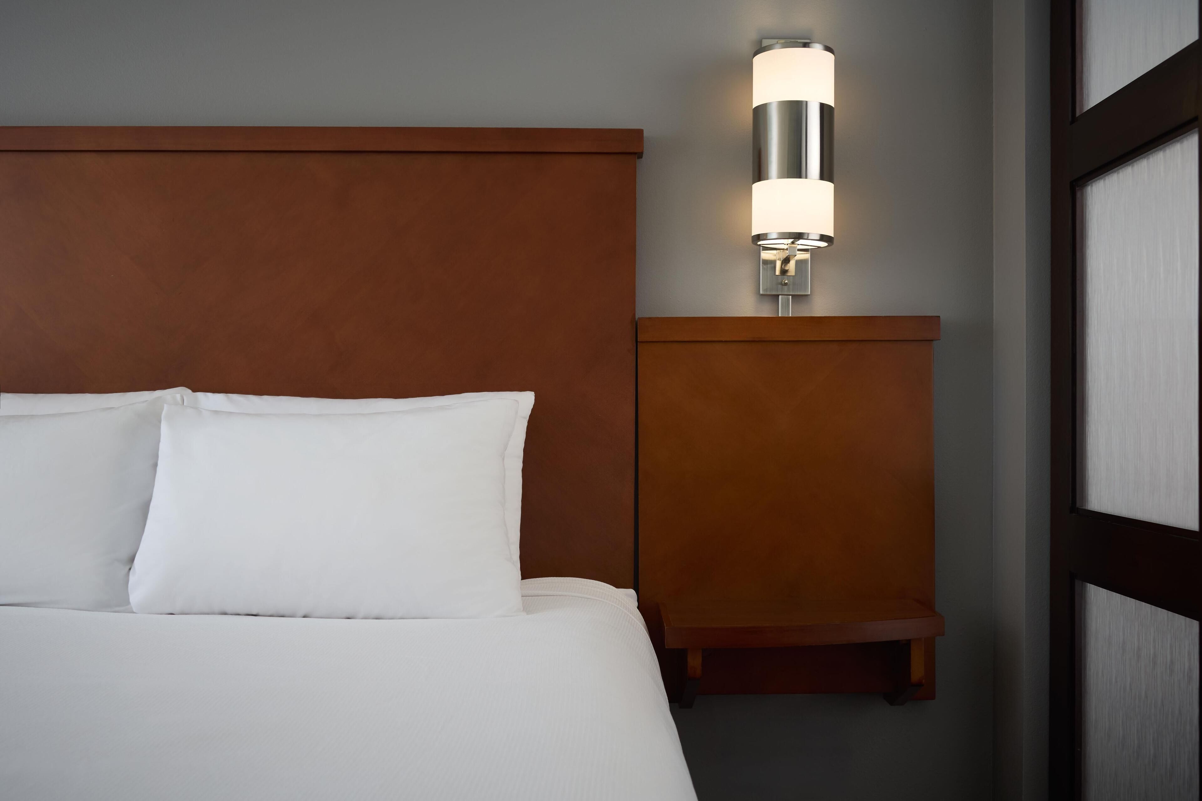 Una cama bien hecha con sábanas y almohadas blancas, un cabecero de madera y una lámpara fijada a la pared.