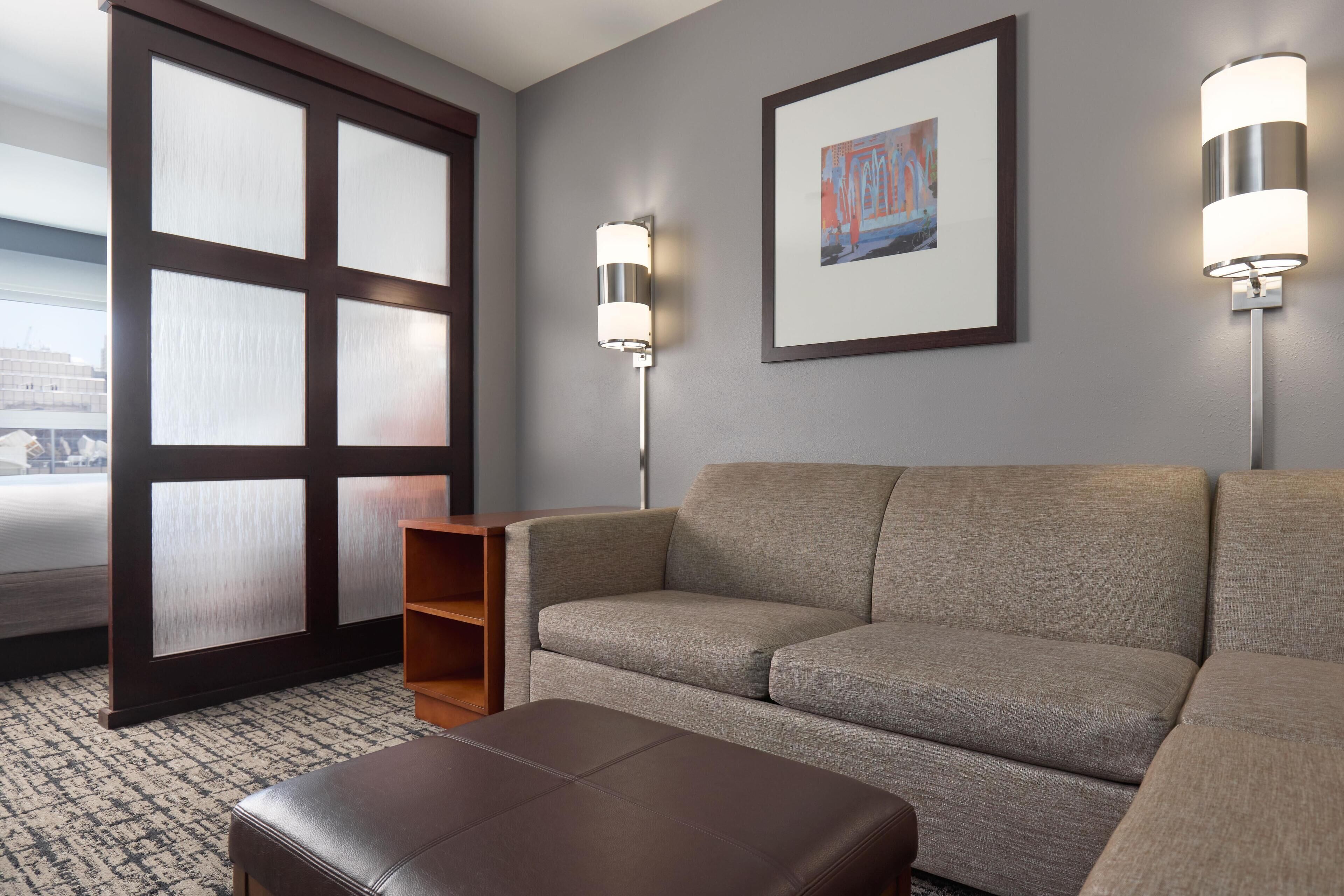 A modern hotel room with a grey sofa, a wooden side table, and two wall-mounted lamps.