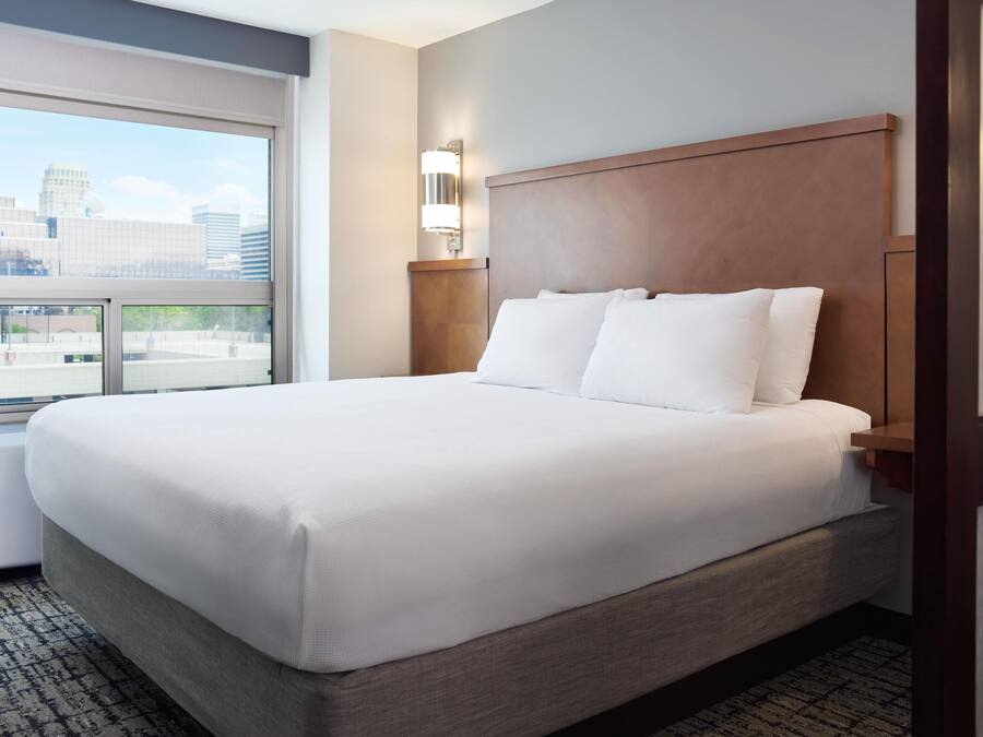 A hotel room with a large bed, a nightstand, a wall-mounted lamp, and a window with a city view.