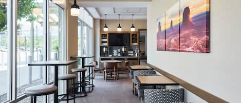 Una cafetería moderna con mesas y taburetes altos, una zona de bar y amplios ventanales con vistas a la calle.