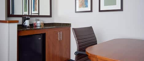 Habitación de hotel con un escritorio de madera, una silla de cuero marrón, refrigerador, cafetera y dos cuadros en la pared.