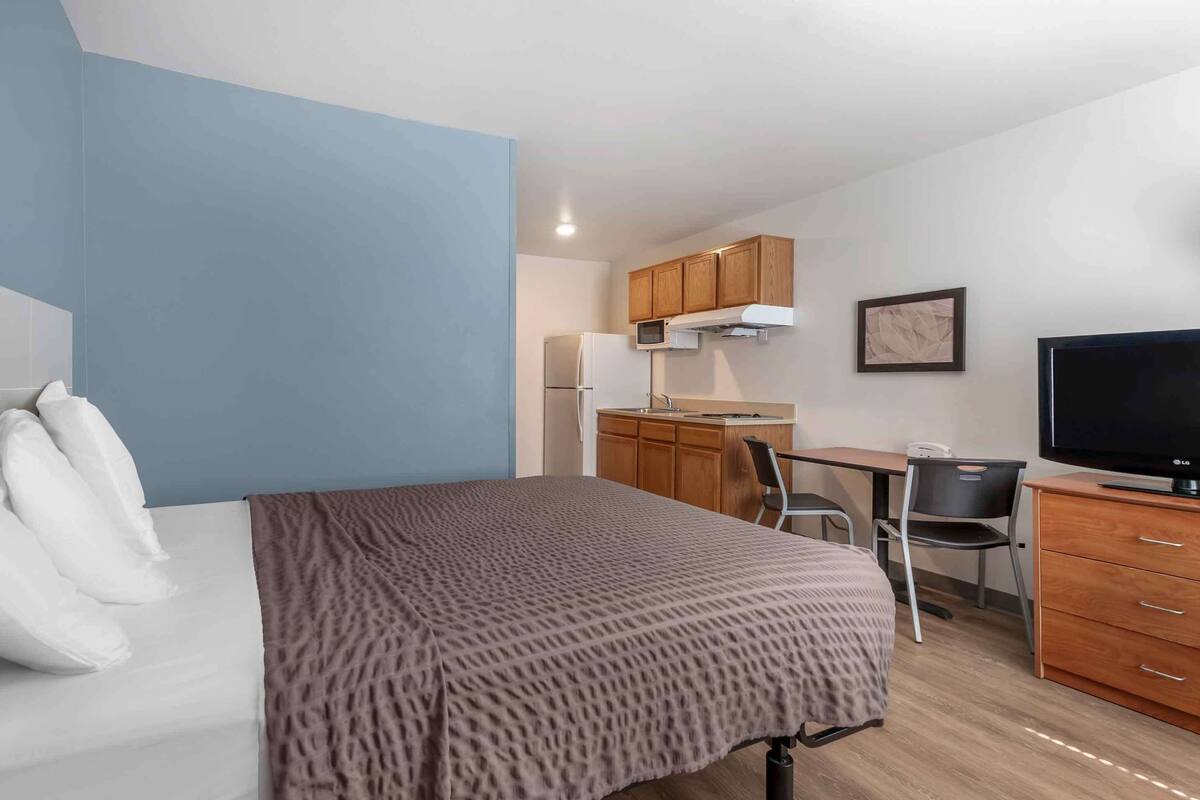 A hotel room with a bed, a desk, a chair, a television, and a kitchenette.