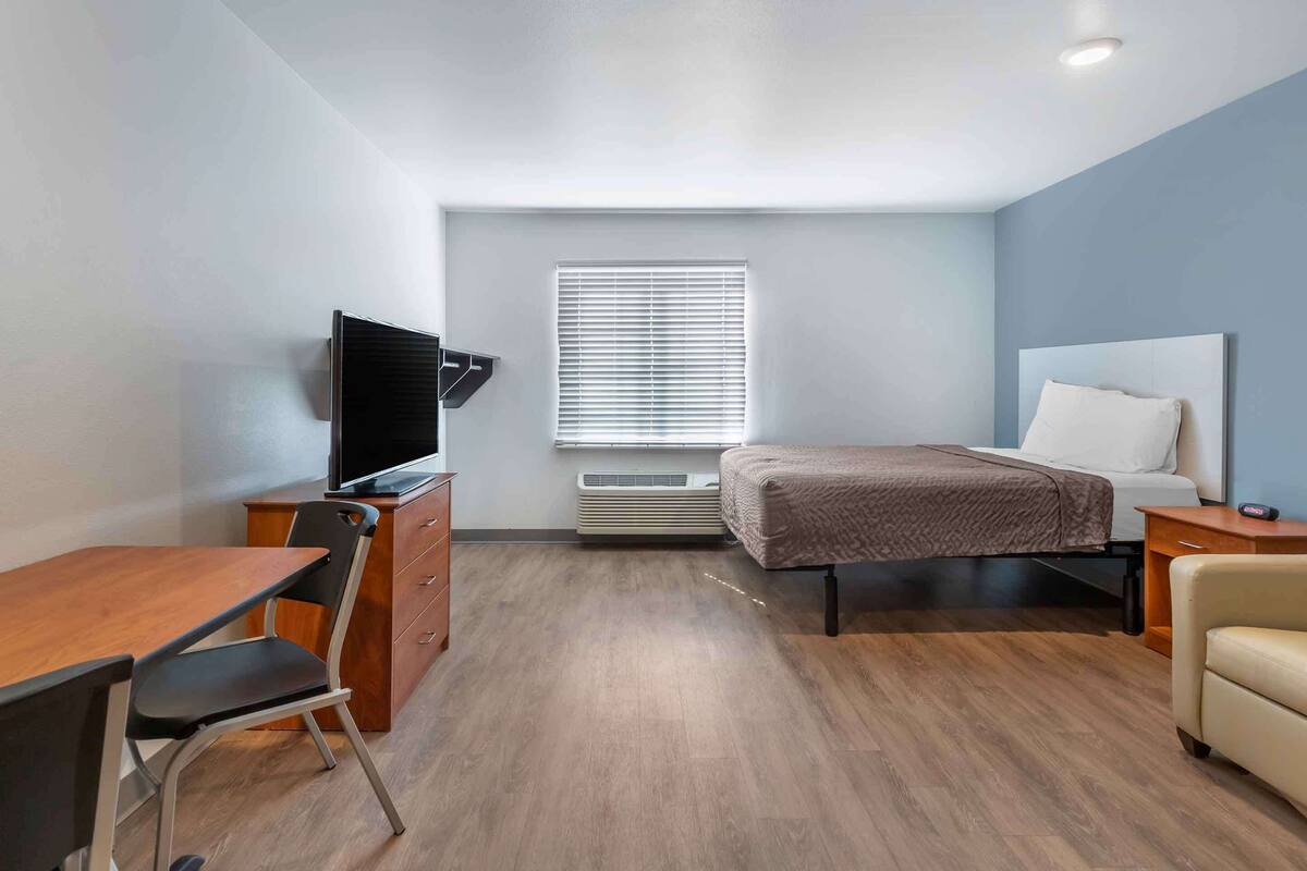 A modern hotel room with a wooden desk, a flat-screen TV, a bed with a white headboard, and a window with blinds.