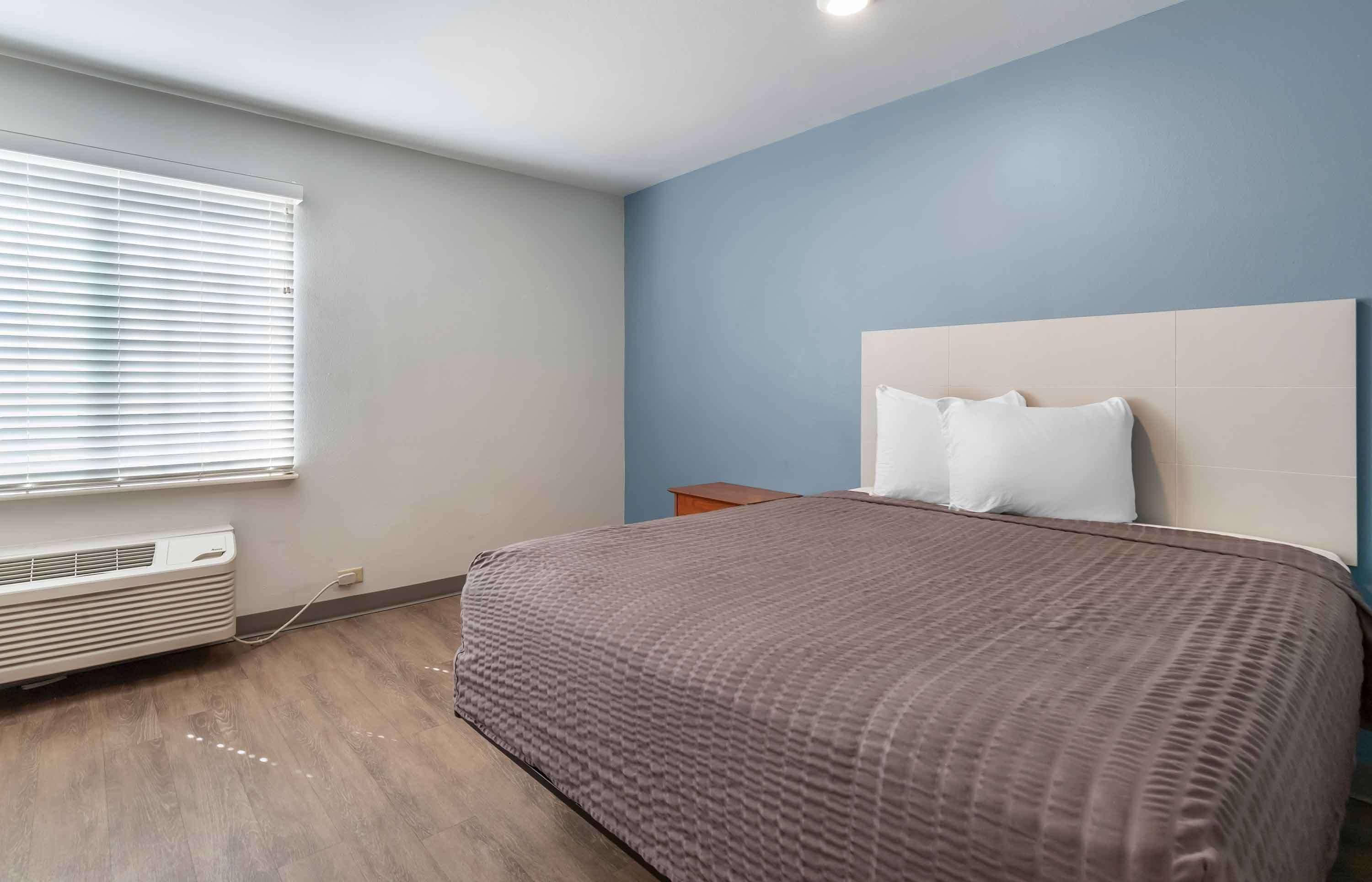 A bedroom with a bed, a nightstand, a window with blinds, and an air conditioning unit.