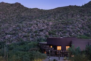 A modern building with a flat roof and large windows, surrounded by desert vegetation and rocky hills.