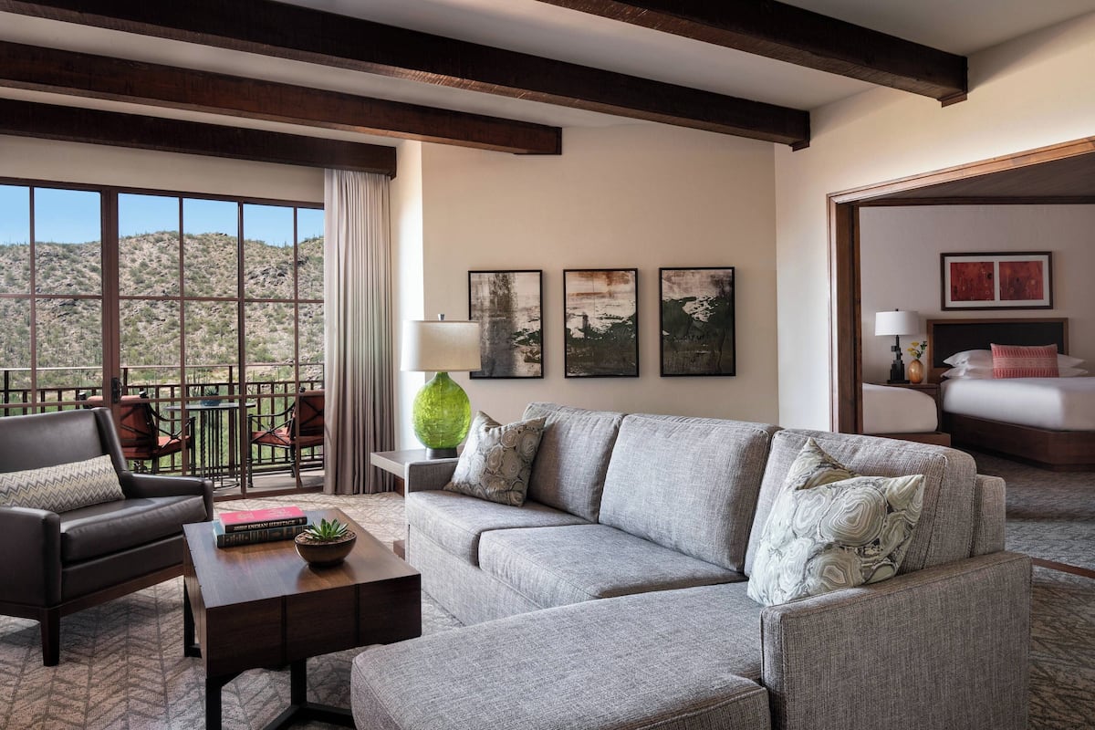 A modern living room with a grey sofa, a brown leather armchair, and a coffee table. There are framed artworks on the wall and a view of a mountainous landscape through large windows.