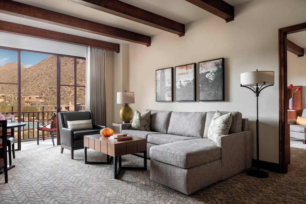 A modern living room with a sectional sofa, a coffee table, and a floor lamp. There is a large window offering a view of a mountainous landscape.