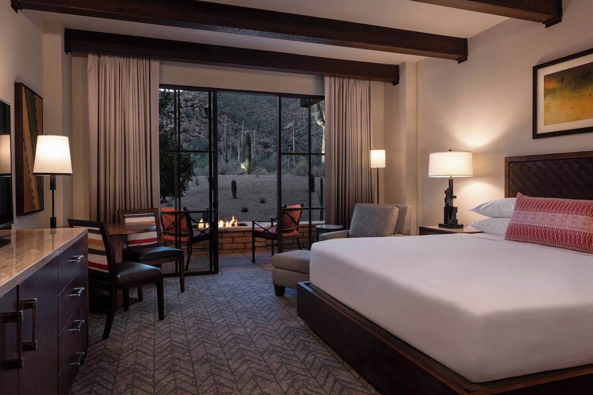 A hotel room with a large bed, two bedside tables with lamps, a desk with a chair, a small table with chairs, and a fireplace.