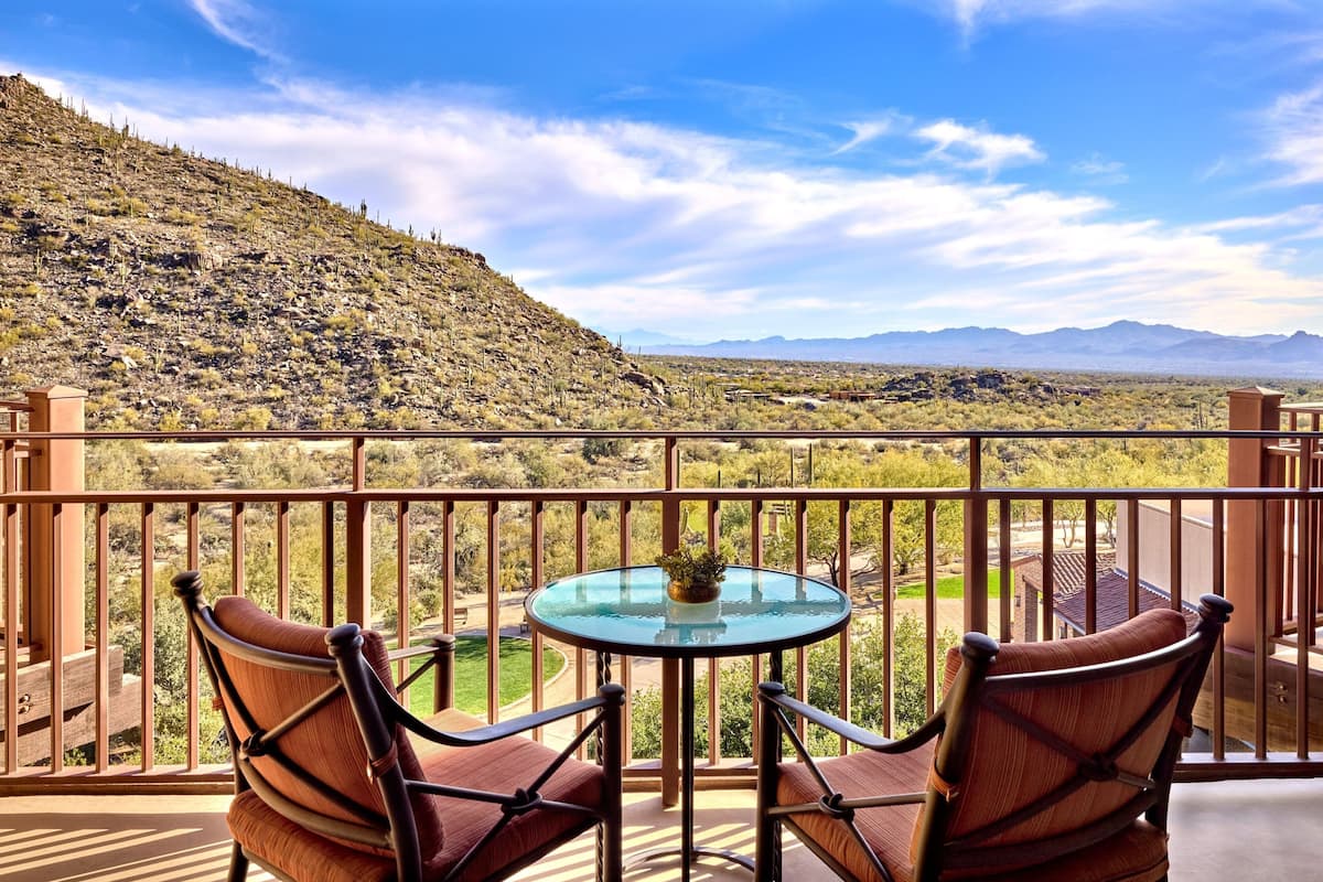 A balcony with two chairs and a glass table, overlooking a mountainous landscape.