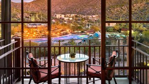 A balcony with a glass table and chairs, overlooking a desert landscape with cacti and buildings.