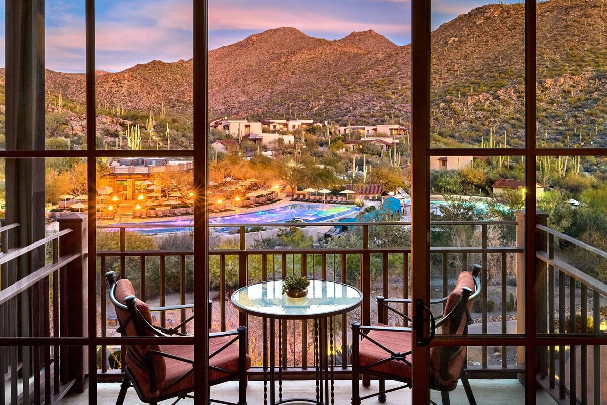 A balcony with a glass table and chairs, overlooking a desert landscape with cacti and buildings.