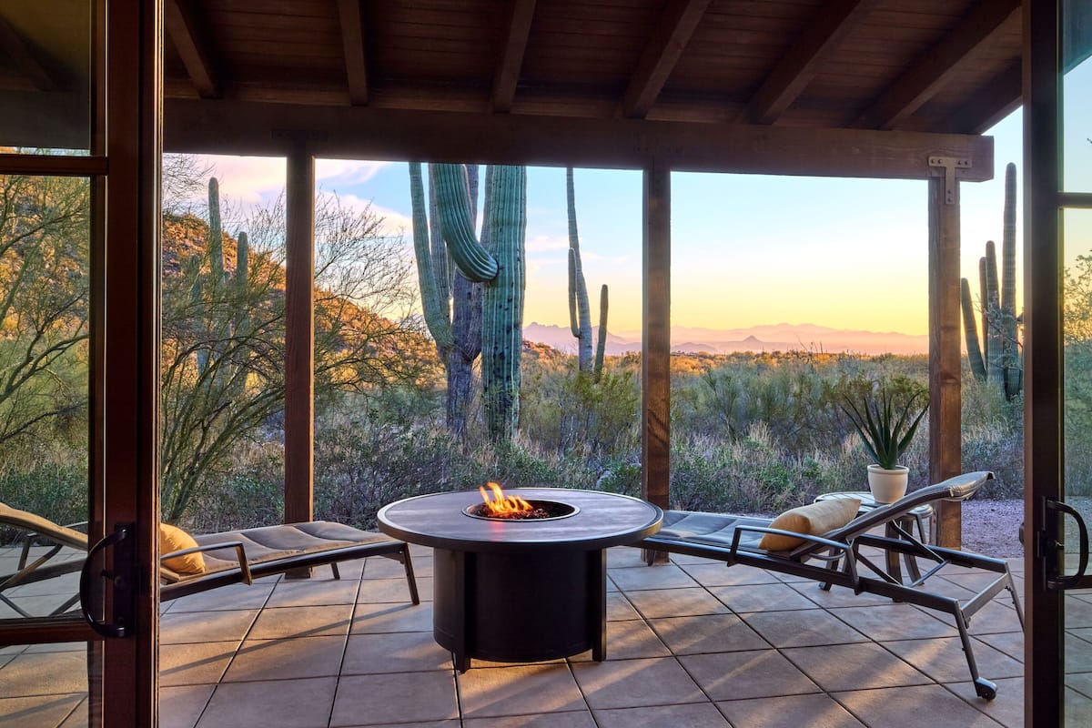 A covered patio with a fire pit, lounge chairs, and a view of desert cacti and mountains.