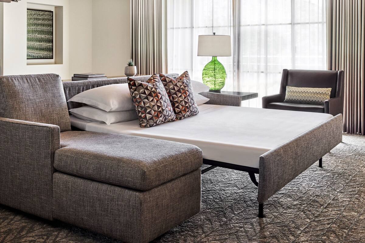 A hotel room with a large bed, a grey upholstered sofa, a small side table with a lamp, and a chair with a cushion.