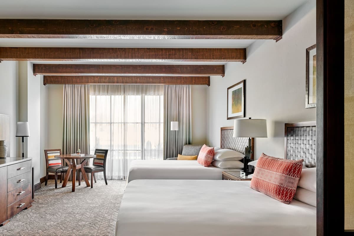 A hotel room with two beds, a dining area with chairs, a desk, and a window with curtains.