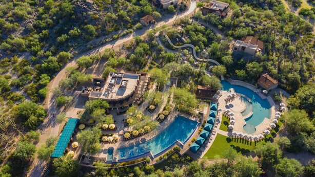 An aerial view of a resort with a large swimming pool, multiple guest areas, and surrounding greenery.