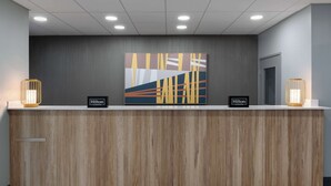 Een receptiebalie van een hotel met een modern ontwerp, met een houten paneel ervoor, een groot abstract kunstwerk erboven en twee verlichte wandlampen aan weerszijden.