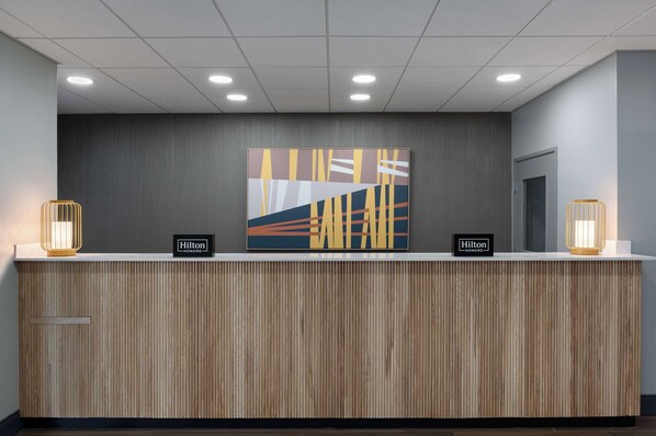 Un mostrador de recepción de hotel con diseño moderno, con un panel frontal de madera, una gran obra de arte abstracta encima de la barra y dos apliques de pared iluminados a cada lado.