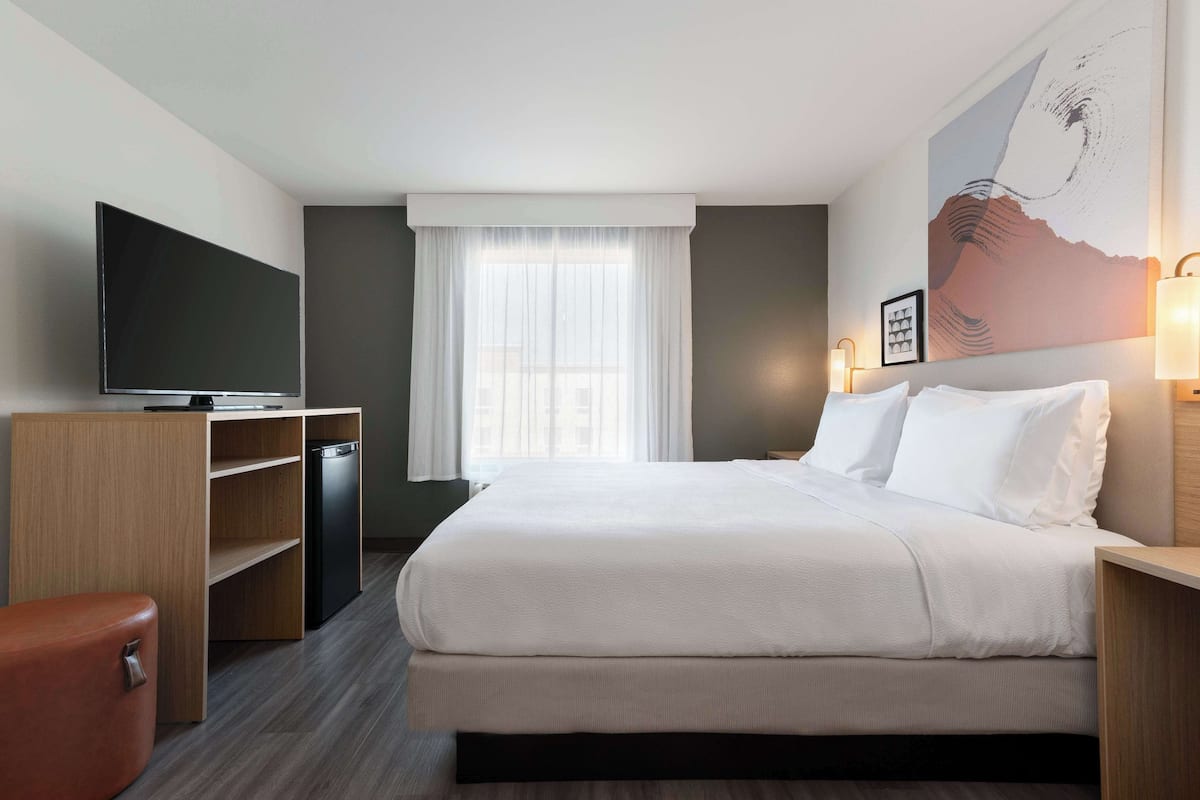 Habitación de hotel con cama, televisor de pantalla plana, escritorio de madera, silla y ventana con cortinas ligeras.