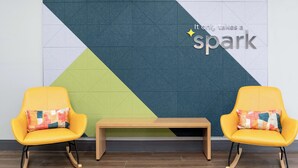 Twee gele stoelen met kleurrijke kussens, een houten bank en een muur met geometrische patronen en de tekst 'It only takes a spark'.