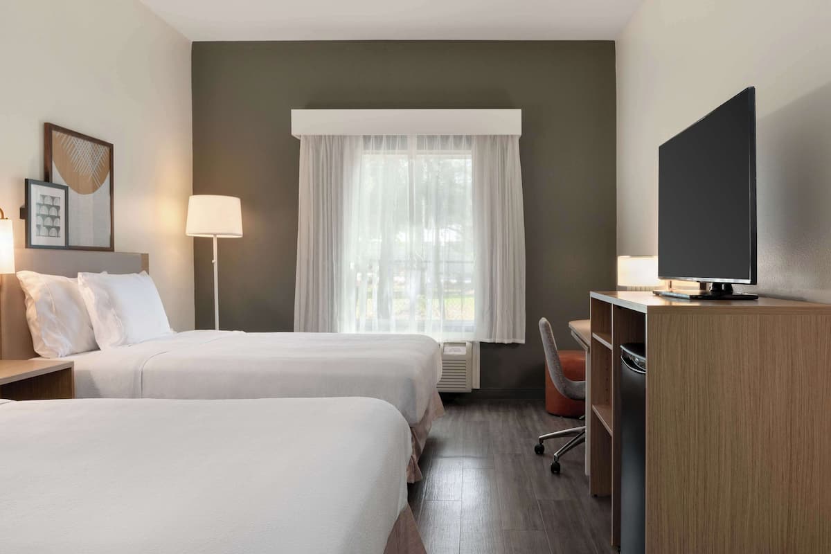 Habitación de hotel con dos camas, un escritorio con televisor, una lámpara y una ventana con cortinas translúcidas.