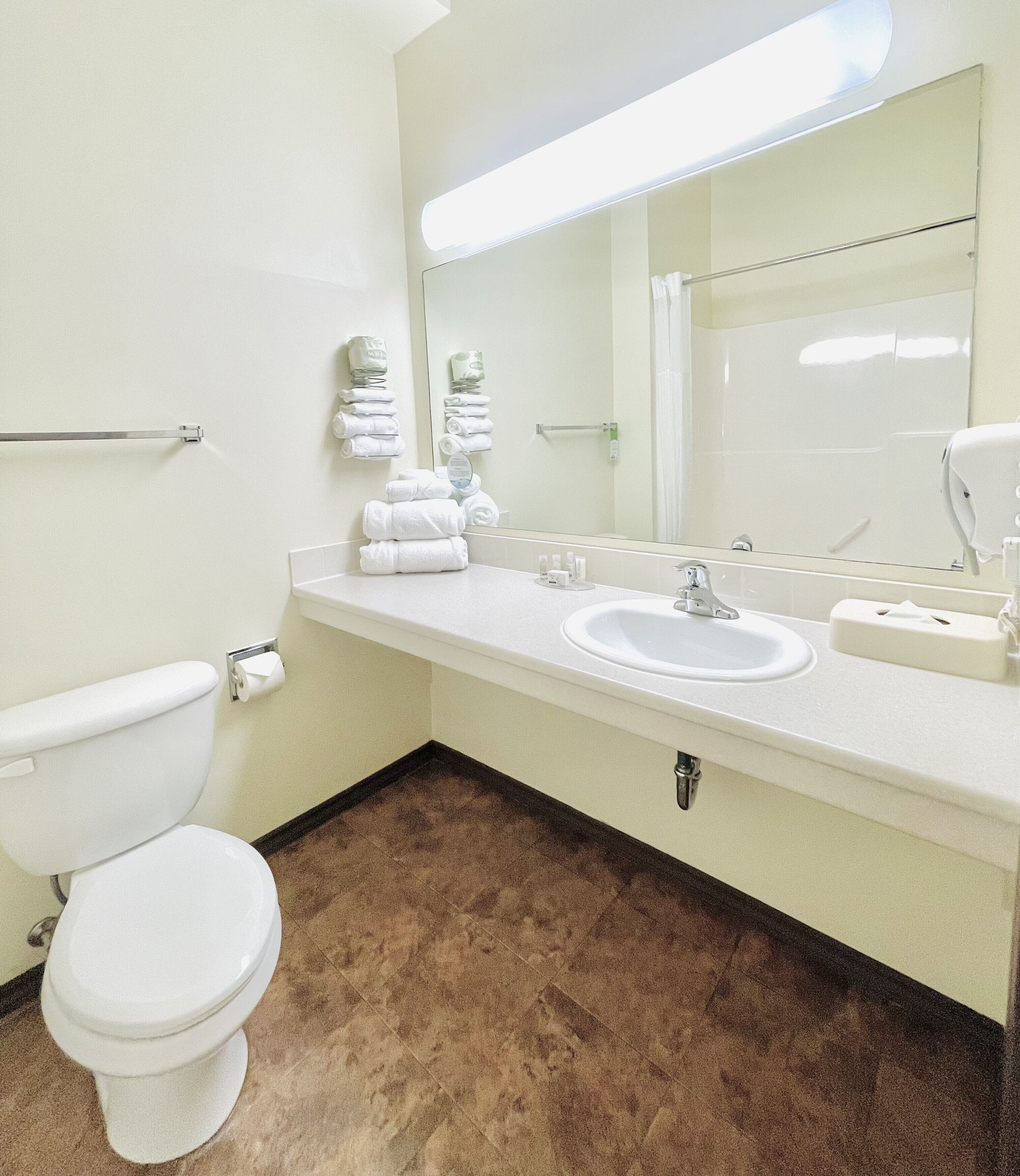 A bathroom with a white toilet, a sink, and a mirror. There are towels stacked on the wall and a soap dispenser on the counter.