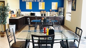 Un coin petit-déjeuner avec une table et des chaises en verre, un mur bleu orné d’affiches motivantes, et un espace cuisine en arrière-plan.