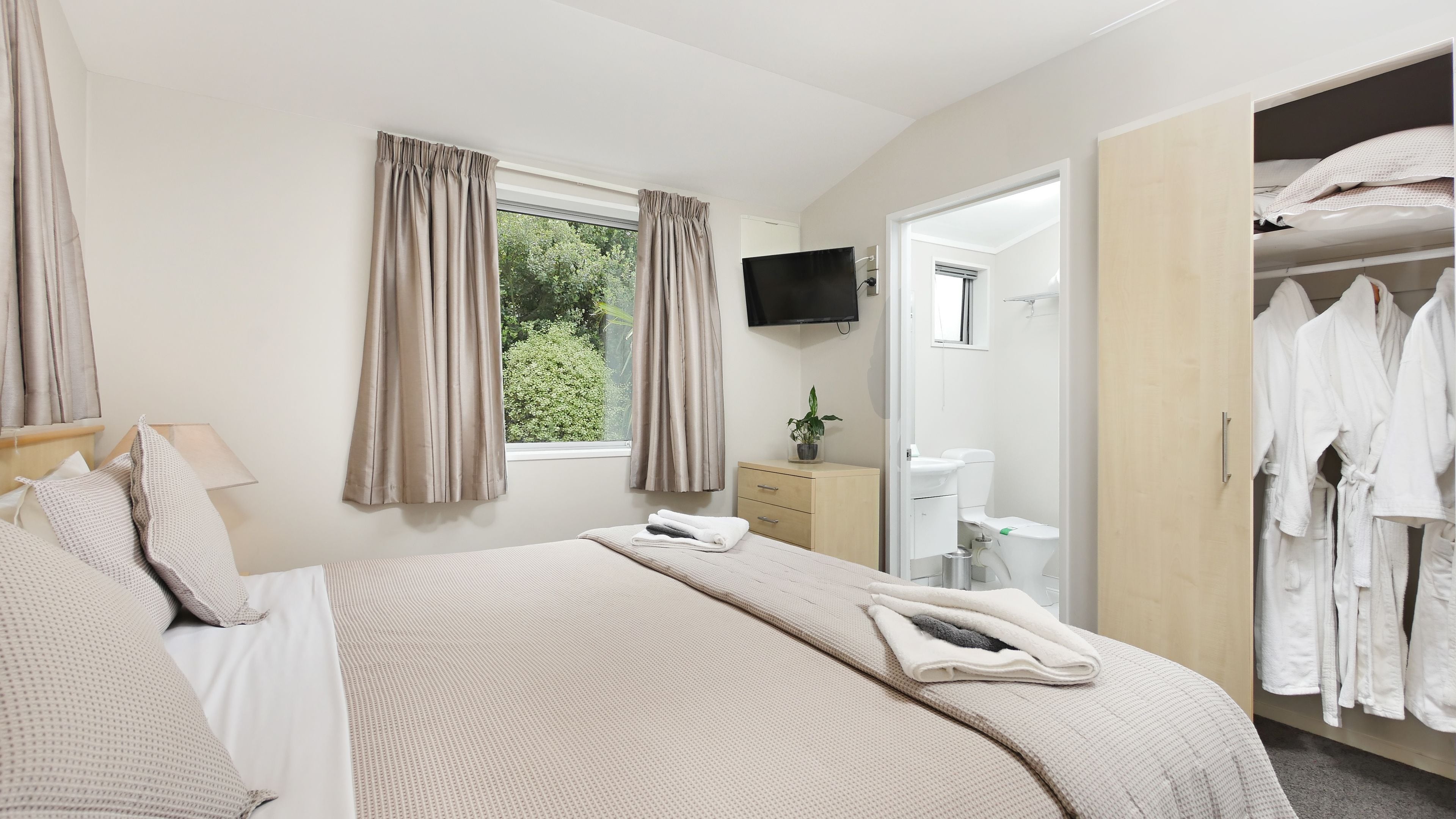 Una habitación de hotel con cama, televisión, ventana con cortinas, baño con inodoro y lavabo, y armario con batas.