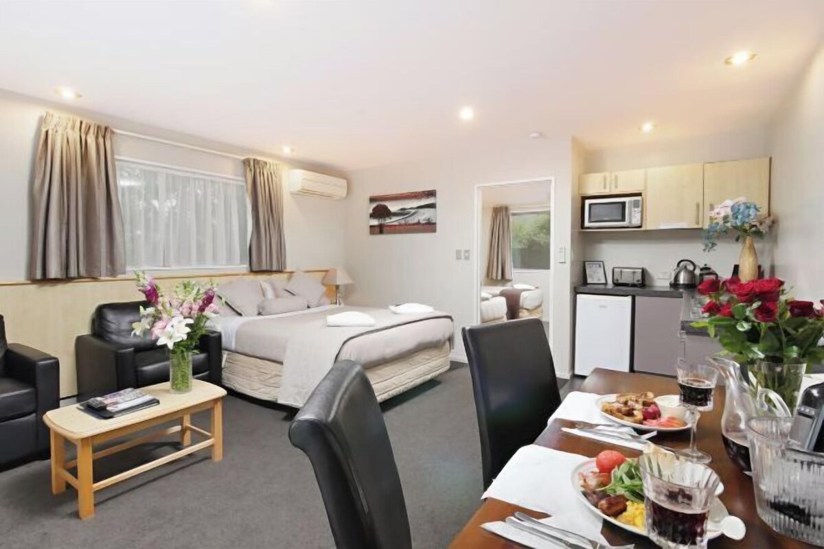 A hotel room with a bed, a small kitchenette, a dining table with breakfast, and a living area with a sofa and coffee table.