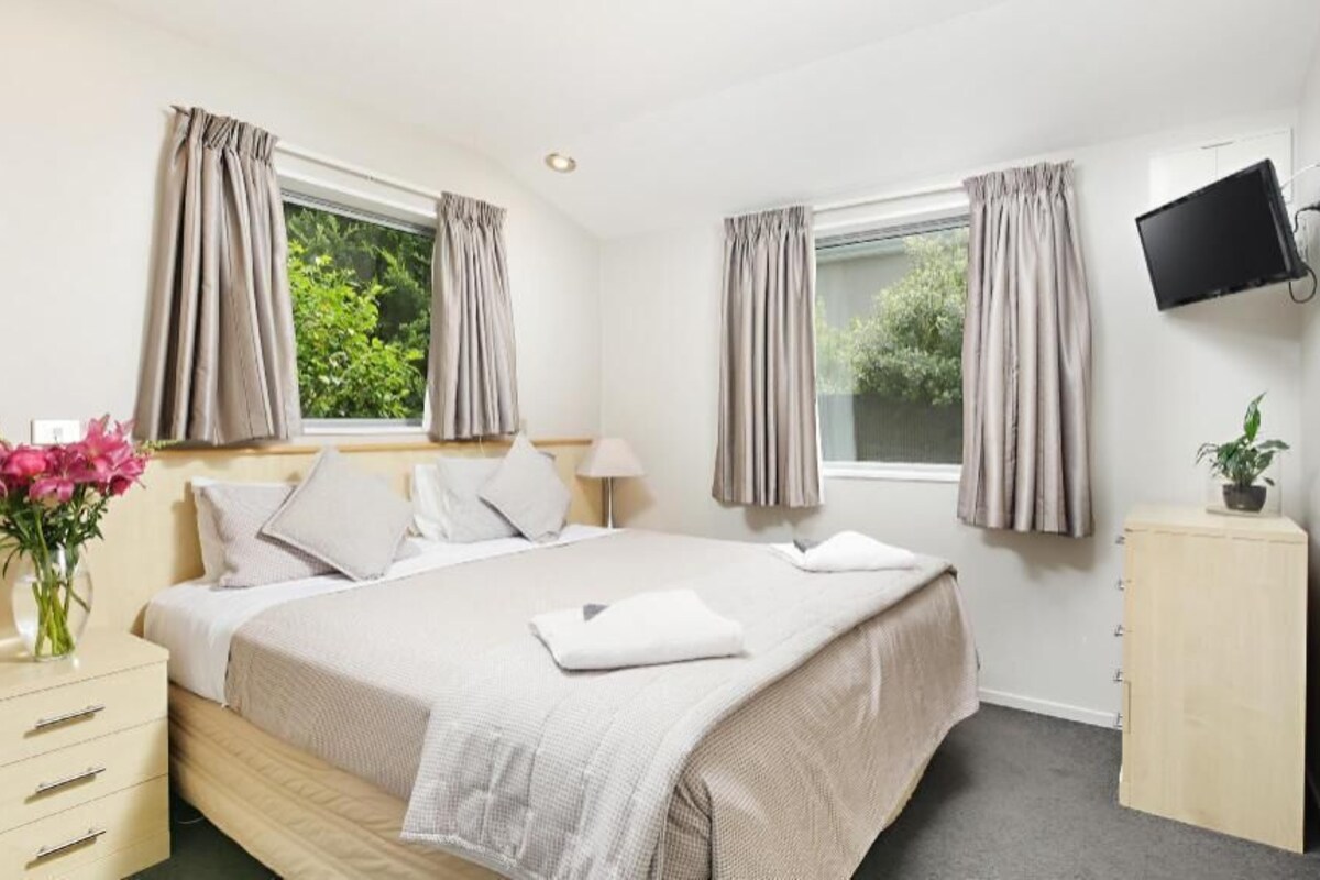 A hotel room with a bed, bedside tables, a TV, and windows with curtains.