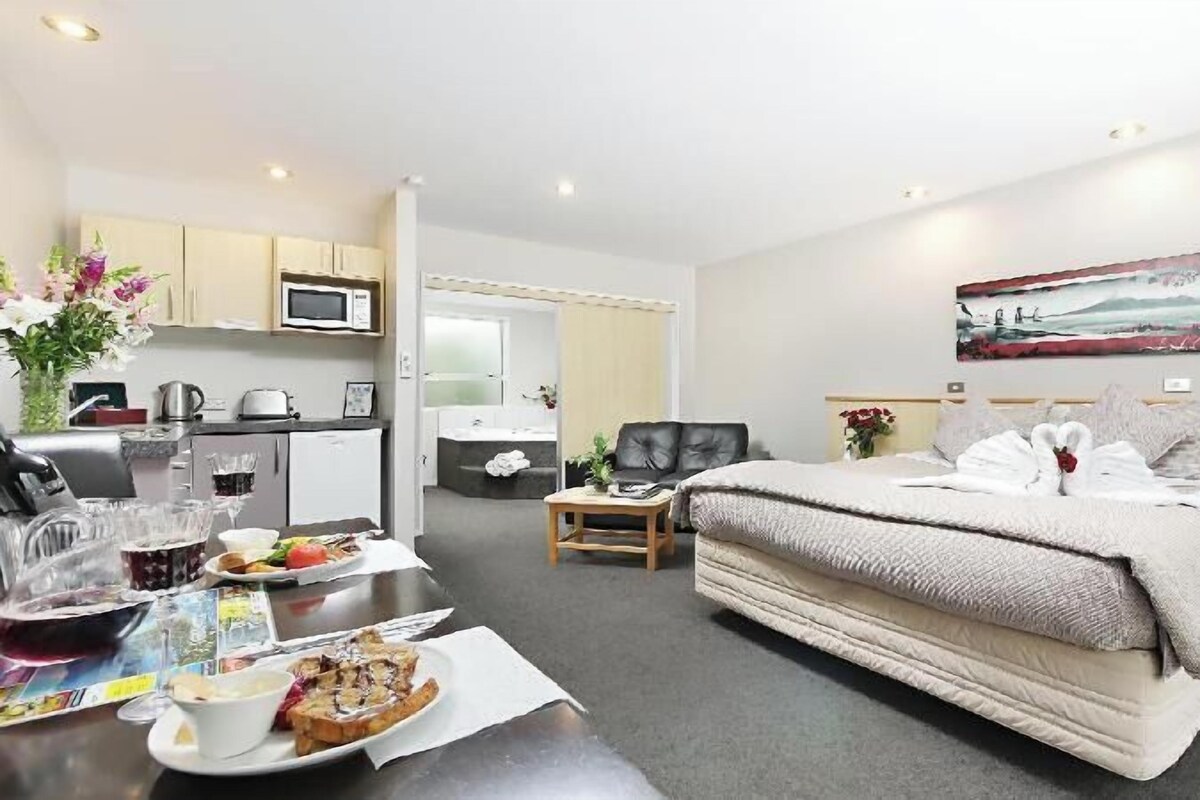 A hotel room with a bed, a dining table with breakfast, a kitchen area, and a bathroom.