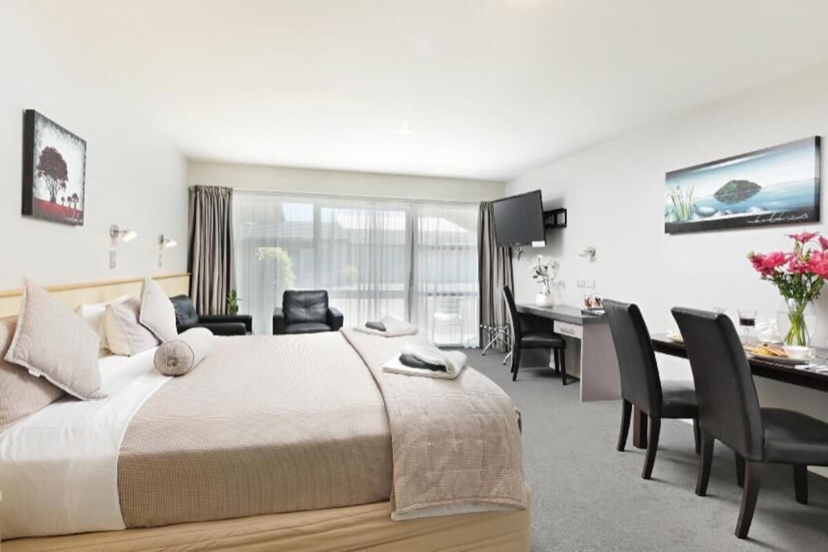 A hotel room with a bed, desk, chairs, a TV, and large windows.