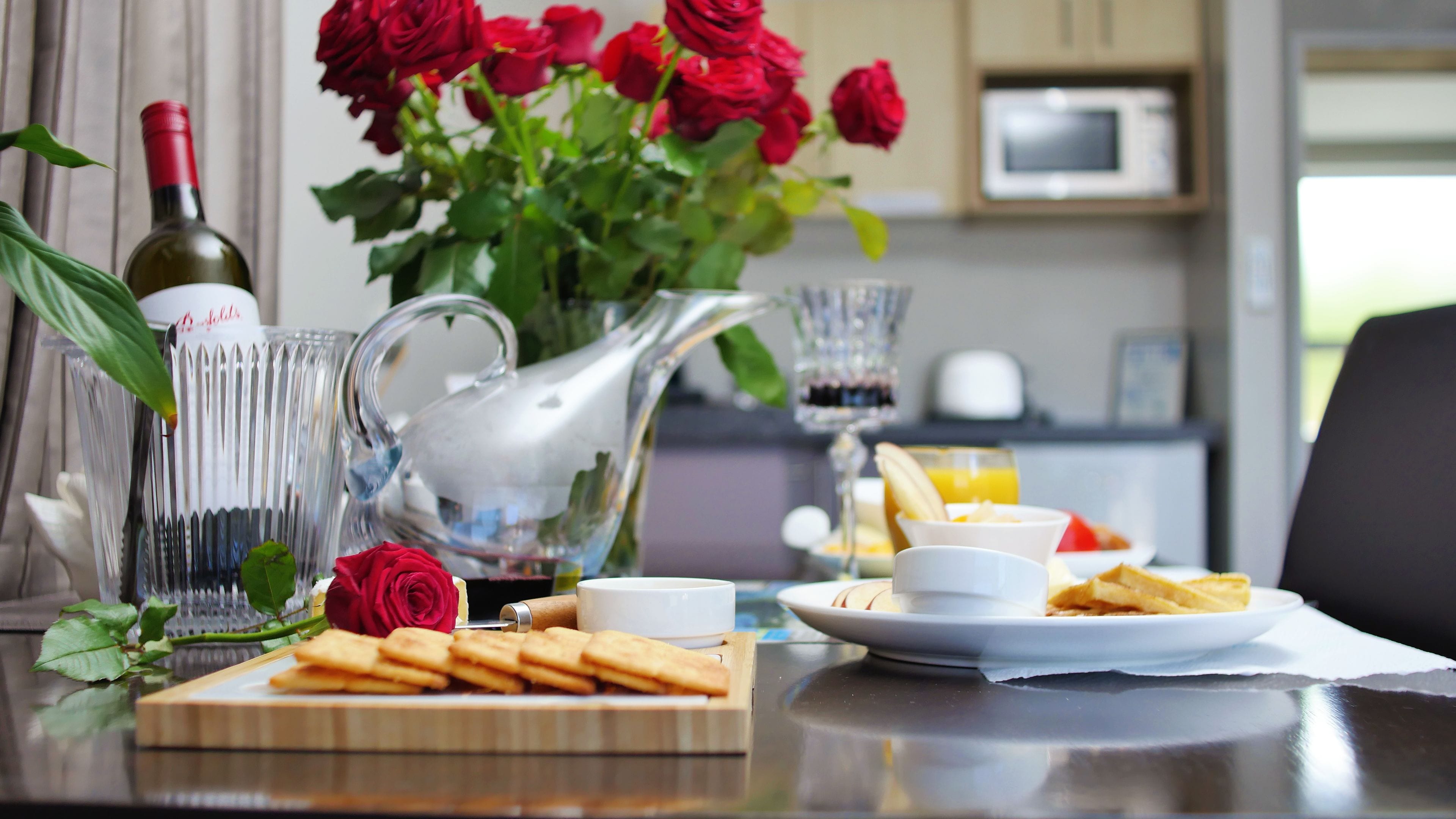 A breakfast table with a bottle of wine, a pitcher, a vase with red roses, a plate of pastries, and a glass of orange juice.