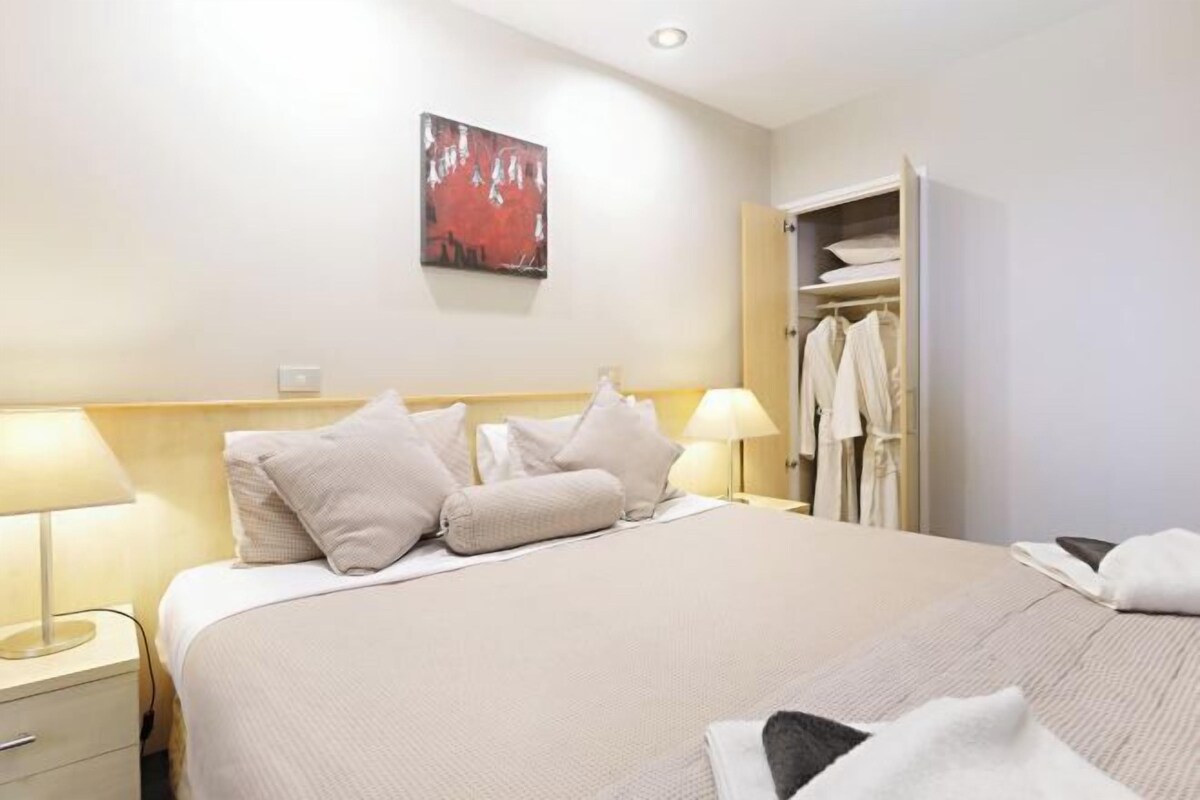 A hotel room with a bed, bedside lamps, a wardrobe, and a painting on the wall.