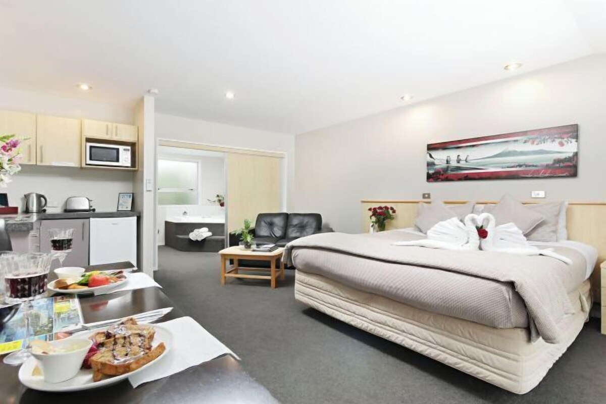 A hotel room with a bed, a desk with breakfast, a microwave, and a painting on the wall.