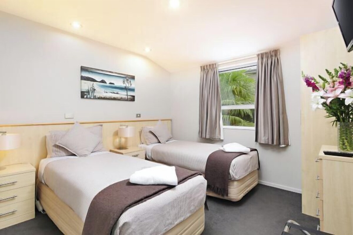 A hotel room with two beds, a flat-screen TV, a window with curtains, a painting on the wall, and a vase with flowers.