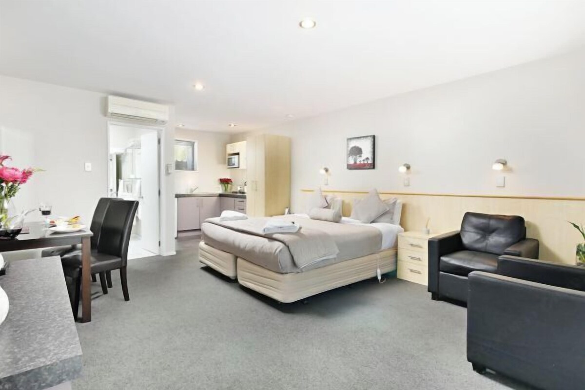 A modern hotel room with a bed, a desk, a chair, a sofa, and a small table with a vase of flowers.