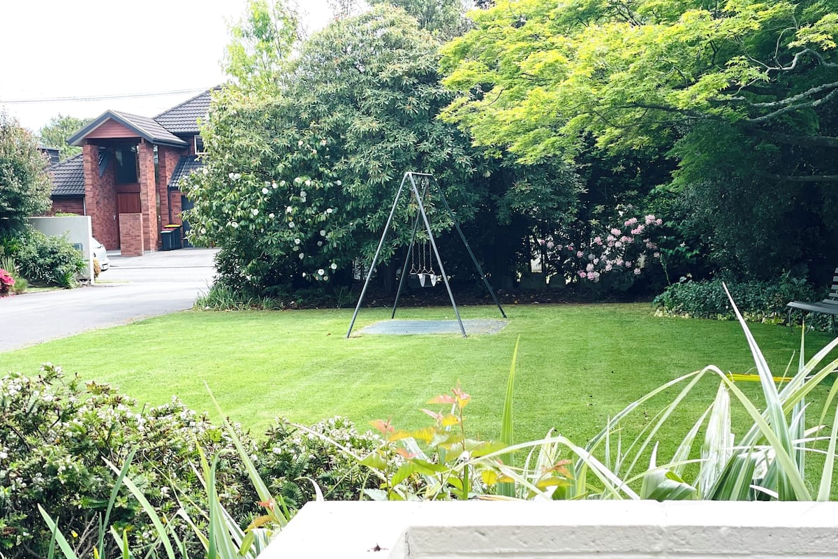 A well-maintained lawn with a swing set, surrounded by lush greenery and a brick building in the background.