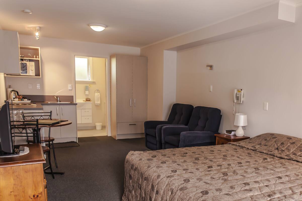A compact hotel room with a bed, a desk with a computer, a chair, a sofa, and a small kitchenette.