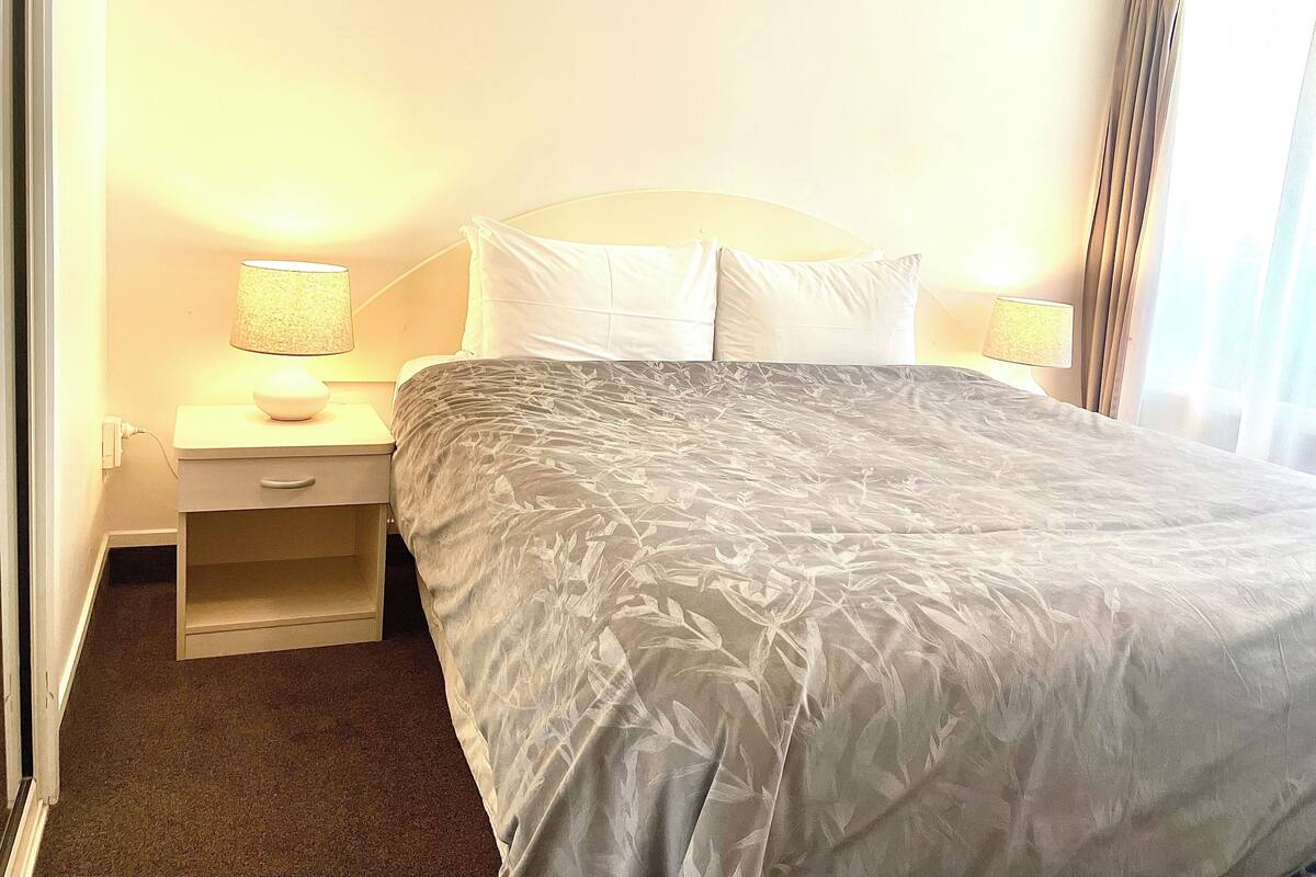 A hotel room with a bed, two bedside lamps, a nightstand, and a small drawer.