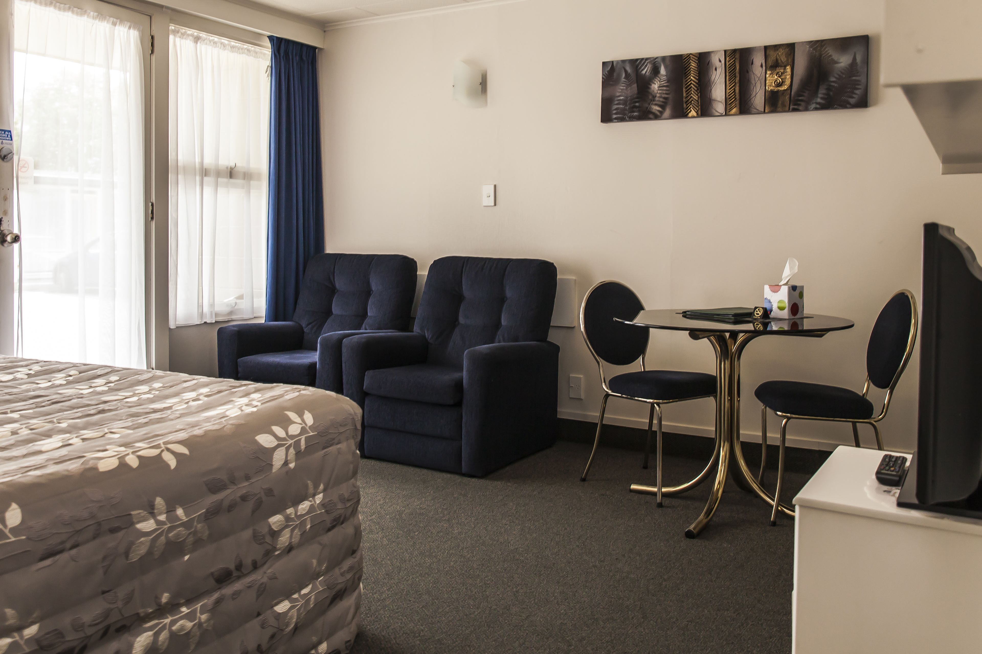 A hotel room with a bed, two armchairs, a small table with a remote control, and a wall-mounted artwork.