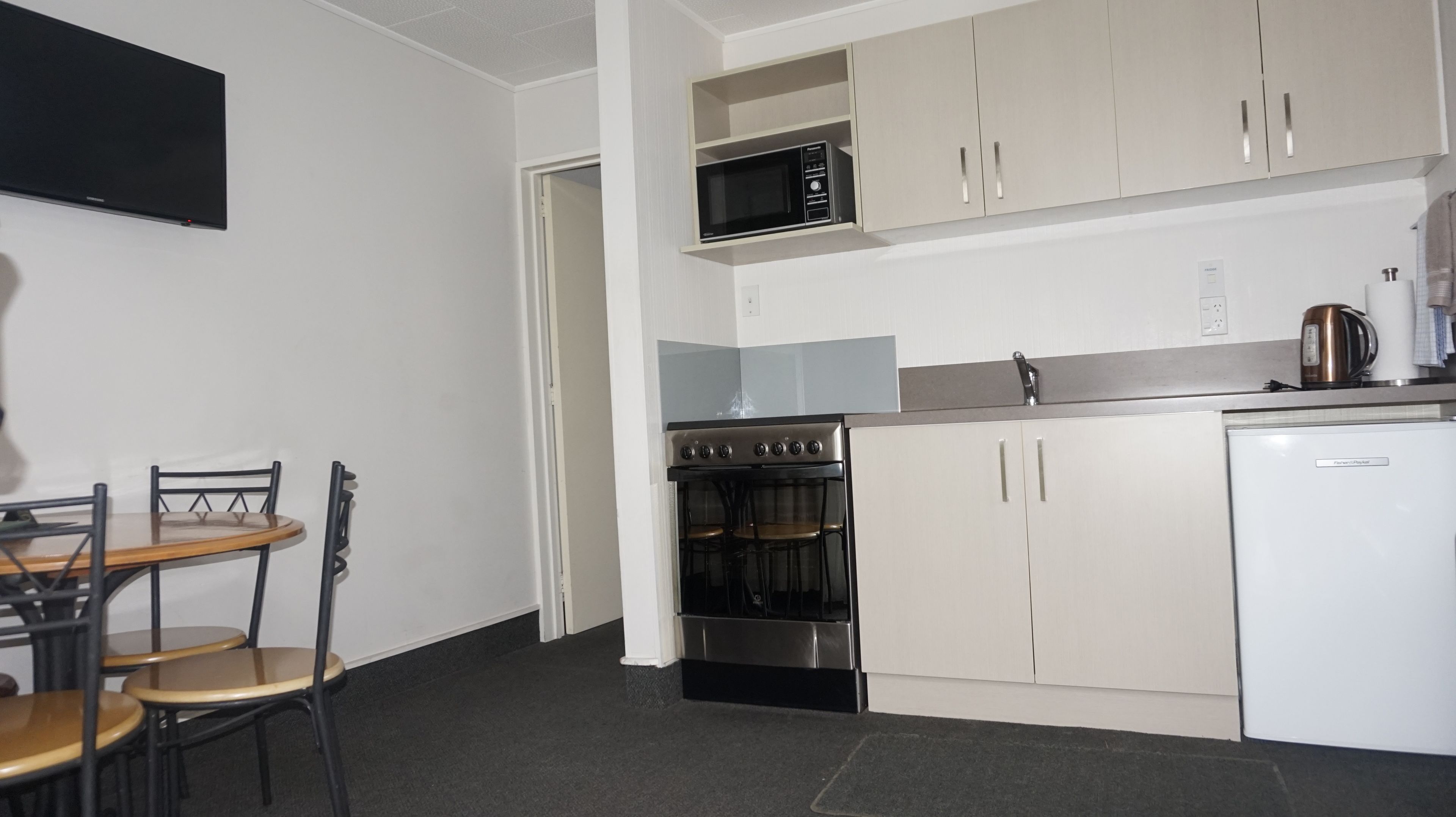 A compact kitchen with a microwave, oven, and a white refrigerator. There is a dining table with chairs and a TV mounted on the wall.