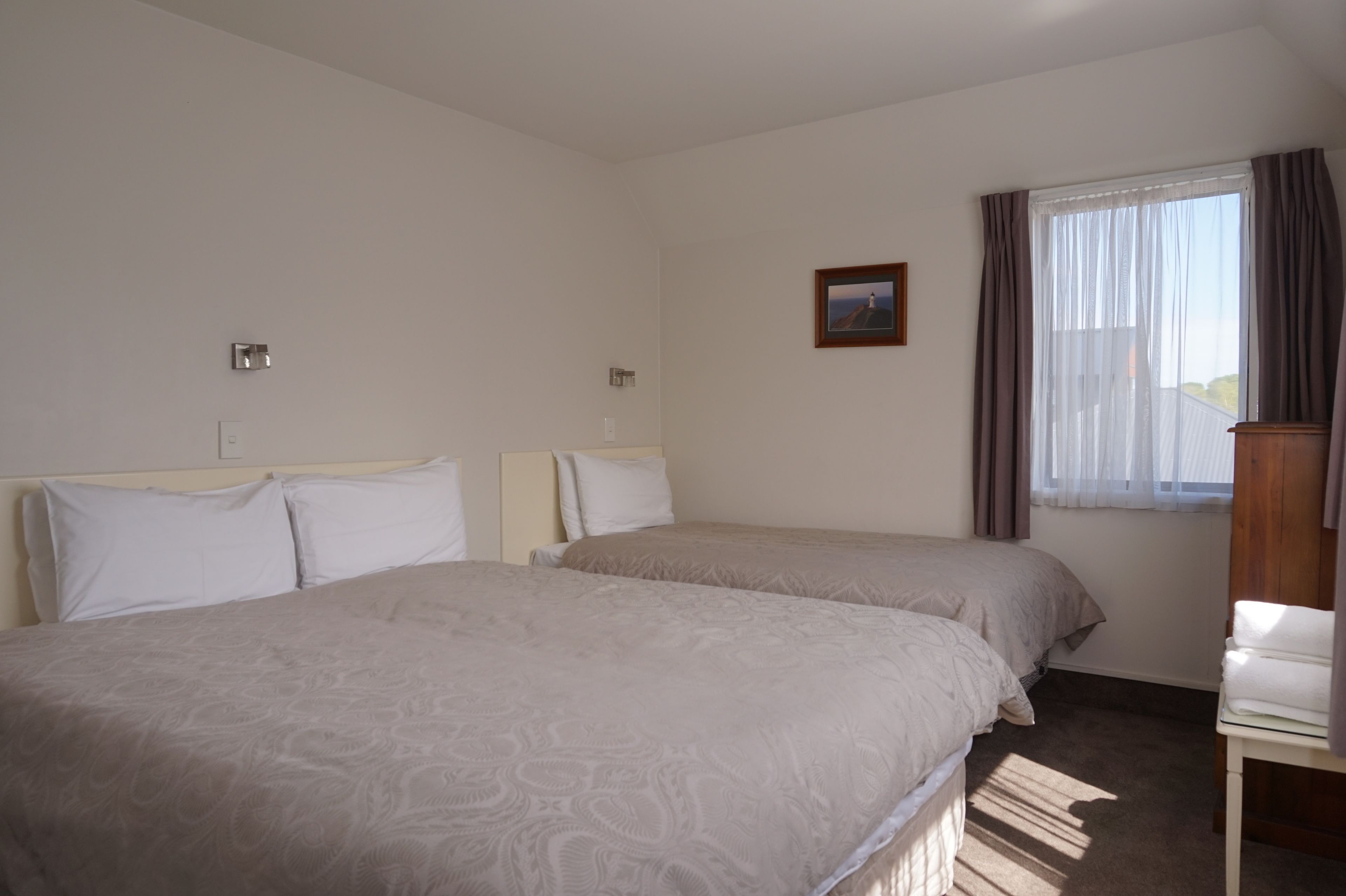 Una habitación de hotel con dos camas, una ventana pequeña con cortinas, un cuadro en la pared y una mesita de noche de madera.