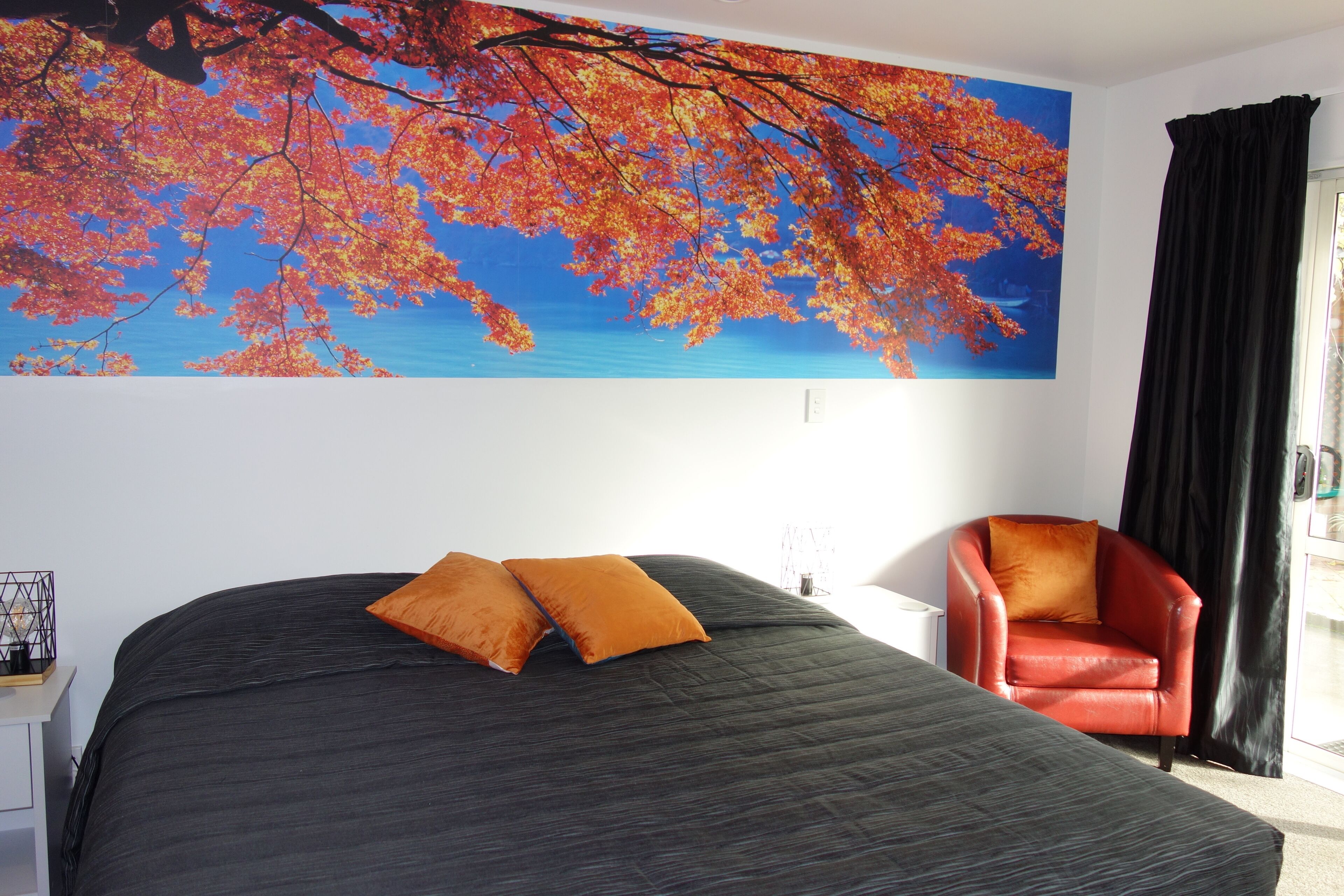 A bedroom with a bed, two orange pillows, a red chair, and a large autumn leaf mural on the wall.