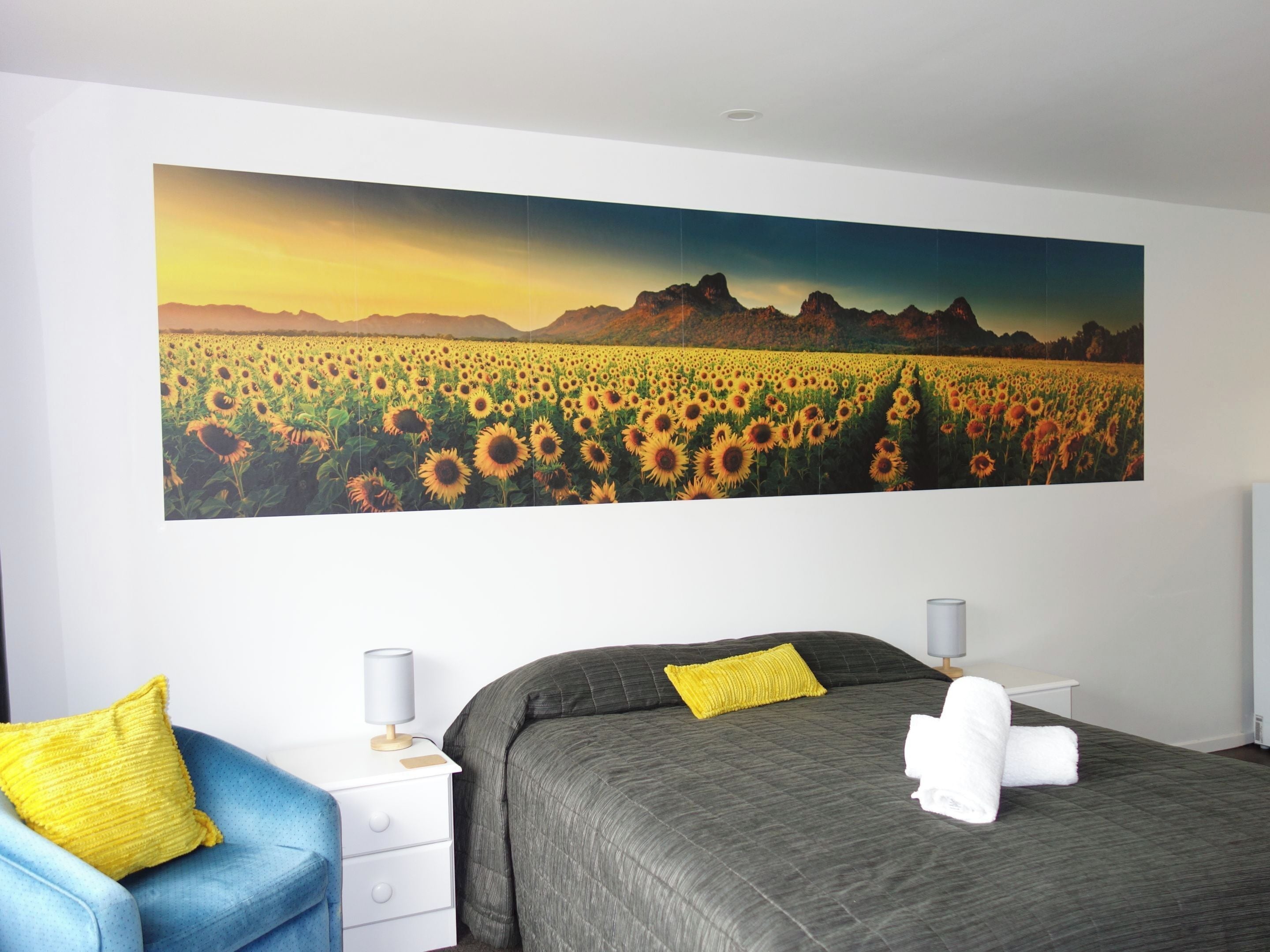 Un dormitorio con un cuadro grande de un campo de girasoles en la pared, una cama con colcha gris y almohada amarilla, una silla azul y una mesita de noche blanca con una lámpara.