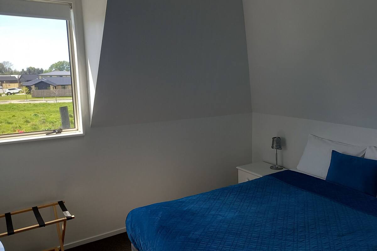 A bedroom with a bed, a nightstand, a lamp, and a window with a view of houses and a grassy area.