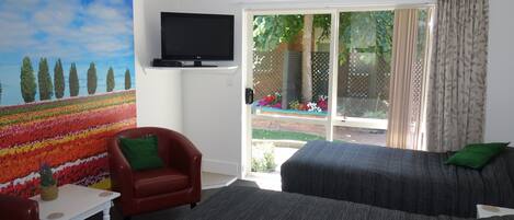 A room with a television mounted on the wall, a red armchair, a grey sofa with green cushions, and a view of a garden through a sliding glass door.