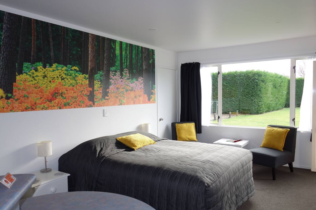 A bedroom with a large bed, two chairs, a desk, and a window with a view of a garden.