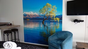 Une chambre d’hôtel dotée d’une grande fresque murale représentant un paysage, d’un téléviseur fixé au mur, d’un coin bar avec des tabourets et d’un espace salon avec une chaise et une petite table.