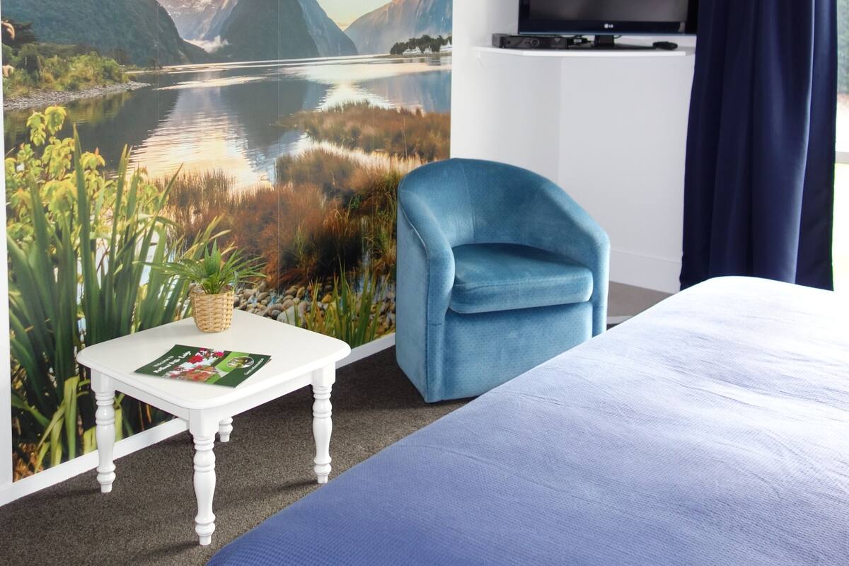 A hotel room with a bed, a blue armchair, a flat-screen TV mounted on the wall, and a scenic mural of a lake and mountains.