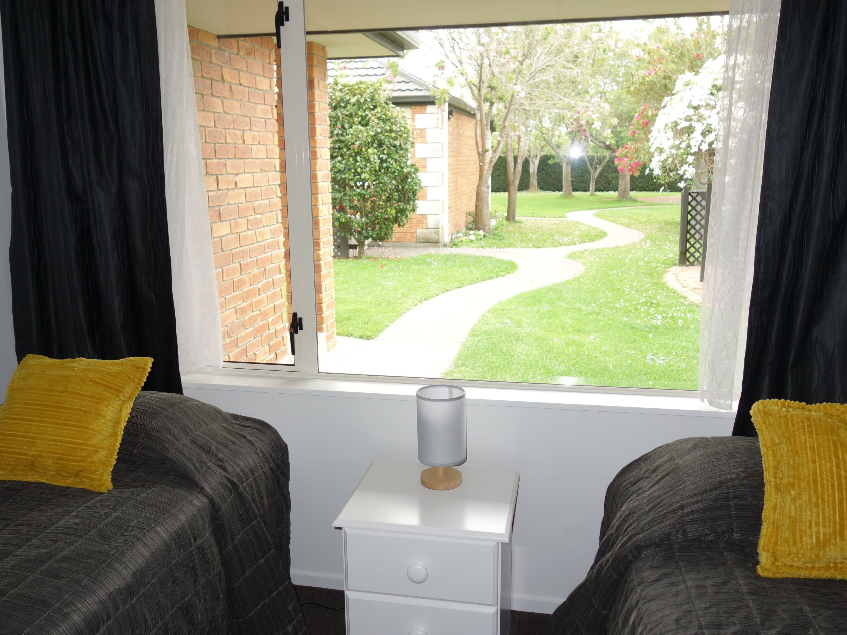 A room with two beds, a small white nightstand, and a window with a view of a garden and a brick wall.